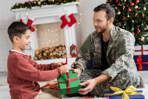 Junge verpackt Geschenkbox in der Nähe von Papa in Tarnung, während er in der Nähe von Kamin mit Weihnachtsdekor sitzt — Stockfoto