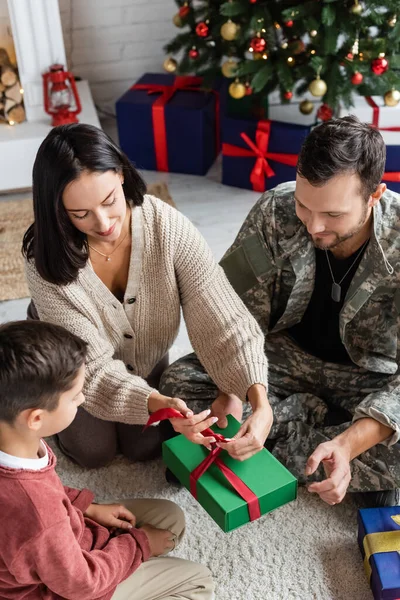 Frau bindet Schleife an Geschenkbox in der Nähe von Sohn und Ehemann in Militäruniform zu Hause — Stockfoto