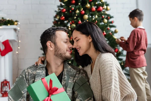 Бородатий військовий чоловік і жінка з закритими очима посміхаються біля подарункової коробки, а син прикрашає ялинку — стокове фото