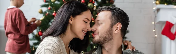 Military man with happy wife smiling face to face with closed eye near son decorating christmas tree, banner — Stock Photo