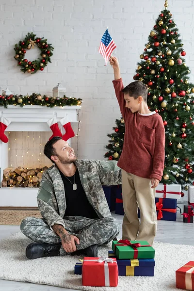 Patriótico chico sosteniendo EE.UU. bandera cerca sonriente padre en militar uniforme sentado cerca de navidad regalos - foto de stock