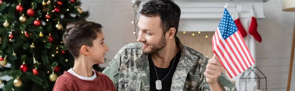 Militar con bandera de EE.UU. sonriendo a hijo feliz cerca del árbol de Navidad, bandera - foto de stock