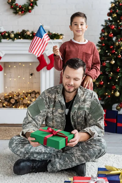 Feliz hombre en uniforme militar sentado con navidad presente cerca de hijo patriótico con bandera de EE.UU. - foto de stock
