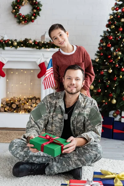 Felice ragazzo con bandiera degli Stati Uniti e l'uomo militare con il regalo di Natale guardando la fotocamera a casa — Foto stock