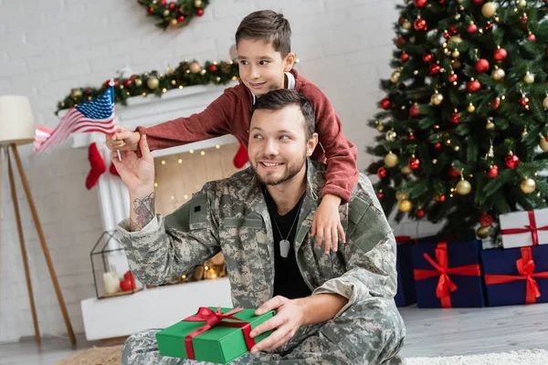 Niño sosteniendo bandera de EE.UU. cerca de padre sonriente en camuflaje sentado con regalo de Navidad - foto de stock