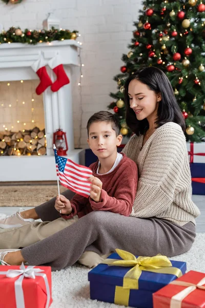 Patriottico ragazzo in possesso di bandiera degli Stati Uniti mentre seduto sul pavimento con sorridente mamma vicino a Natale presenta — Foto stock