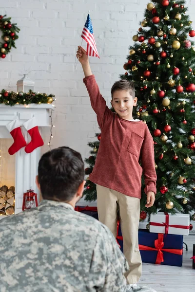 Smiling patriotic boy standing with usa flag near christmas tree and dad in camouflage on blurred foreground — Stock Photo