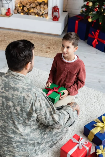 Vue grand angle du garçon souriant assis sur le sol près de papa en camouflage et tenant cadeau de Noël — Photo de stock