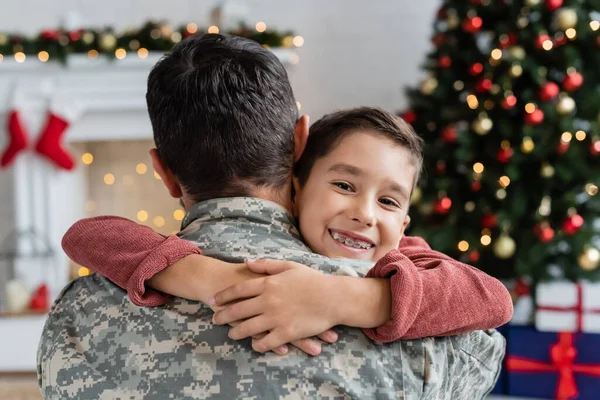 Gioioso ragazzo in denti bretelle abbracciando il padre in uniforme militare e sorridendo alla fotocamera vicino all'albero di Natale offuscato — Foto stock