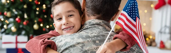 Niño feliz sosteniendo bandera de EE.UU. y abrazando al padre en uniforme militar en casa, pancarta - foto de stock