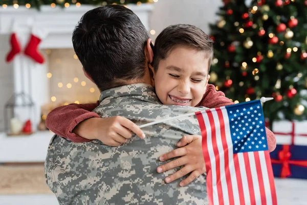 Niño alegre con los ojos cerrados y la bandera de EE.UU. abrazando a papá en camuflaje en casa - foto de stock