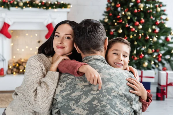 Happy mother and son embracing man in camouflage near blurred christmas tree and decorated fireplace — Stock Photo