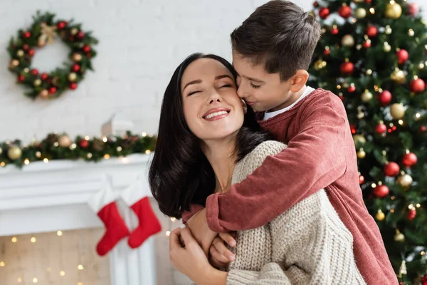 Menino abraçando mãe feliz perto de árvore de Natal borrada em casa — Fotografia de Stock