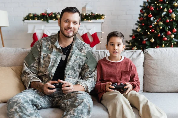 QUIIV, UCRÂNIA - SETEMBRO 21, 2022: homem militar feliz jogando videogame com filho em casa — Fotografia de Stock