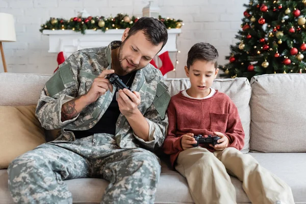 KYIV, UKRAINE - 21 SEPTEMBRE 2022 : Homme militaire souriant regardant joystick tout en jouant à un jeu vidéo avec son fils à la maison — Photo de stock