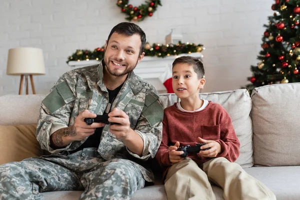 KYIV, UKRAINE - SEPTEMBER 21, 2022: excited boy and father in military uniform gaming with joysticks at home — Stock Photo