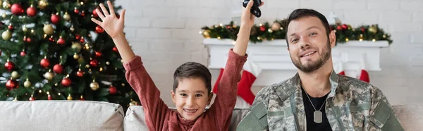 KIEW, UKRAINE - 21. SEPTEMBER 2022: fröhlicher Junge mit erhobenen Händen, Joystick neben lächelndem Vater in Tarnkleidung, Banner — Stockfoto