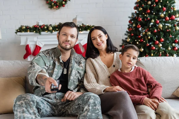 Feliz militar haciendo clic en canales mientras ve la televisión con la familia cerca del árbol de Navidad - foto de stock