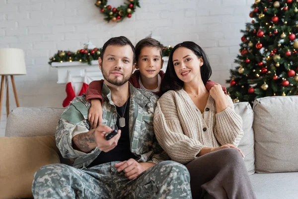 Menino abraçando feliz mãe e pai em camuflagem assistindo tv perto da árvore de natal — Fotografia de Stock