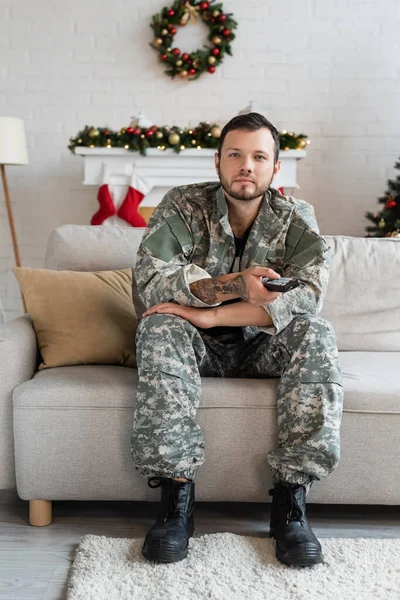 Militar homem com tv controle remoto sentado no sofá na sala de estar com decoração de Natal — Fotografia de Stock