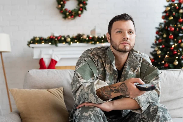 Unrasierter Mann in Tarnkleidung hält Fernbedienung in der Hand, während er zu Hause in der Nähe des verschwommenen Weihnachtsbaums fernsieht — Stockfoto