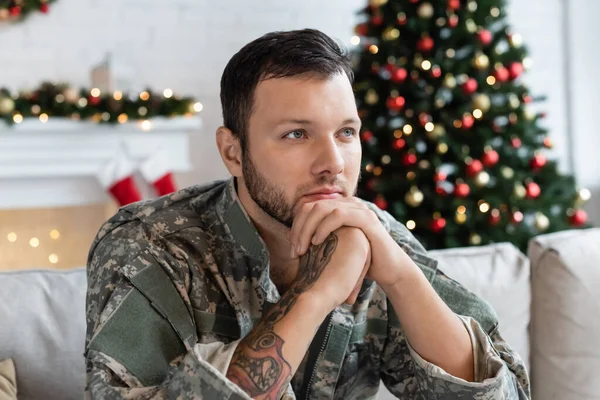 Hombre reflexivo en camuflaje sentado con las manos cerca de la cara y mirando hacia otro lado cerca borrosa árbol de Navidad — Stock Photo