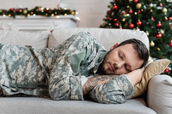 Втомлений бородатий чоловік у військовій формі спить на дивані вдома в різдвяний день — стокове фото