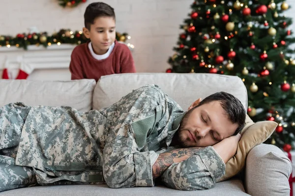 Расплывчатый мальчик смотрит на измученного папу в камуфляже спящего на диване в гостиной с рождественским декором — стоковое фото
