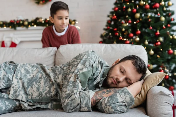 Blurred boy looking at exhausted dad in camouflage sleeping on couch in living room with christmas decoration — Stock Photo