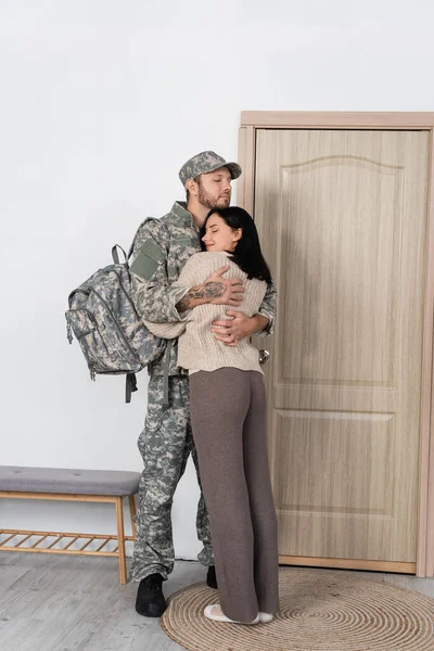 Hombre en uniforme militar abrazando esposa feliz cerca de la puerta de entrada en casa - foto de stock