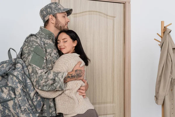 Щаслива жінка з закритими очима обіймає чоловіка у військовій формі біля вхідних дверей вдома — стокове фото