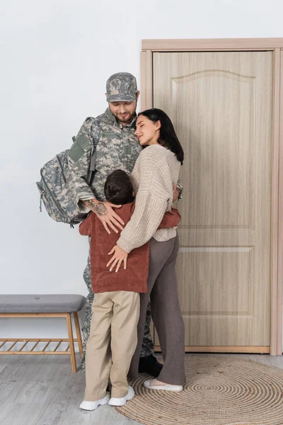 Homem em camuflagem abraçando filho e mulher feliz perto da porta de entrada em casa — Fotografia de Stock