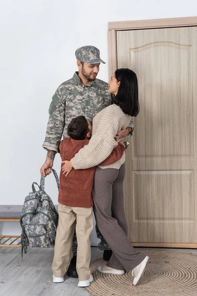 Militar sin afeitar con mochila abrazando esposa e hijo encontrándolo en casa - foto de stock