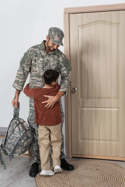 Niño abrazando papá en uniforme militar de pie con mochila cerca de la puerta de entrada en casa - foto de stock