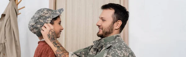 Vue latérale du papa souriant portant un bonnet militaire sur son fils lors de son retour à la maison, bannière — Photo de stock