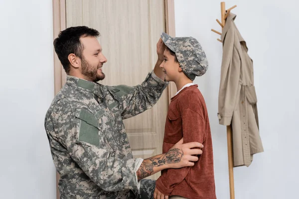 Seitenansicht des fröhlichen Jungen mit Militärmütze neben lächelndem Vater in Tarnkleidung — Stockfoto