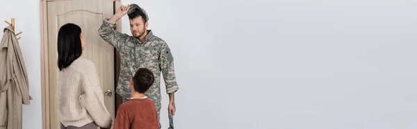 Hombre en camuflaje quitándose la gorra militar cerca de la esposa y el hijo encontrándolo en casa, pancarta - foto de stock