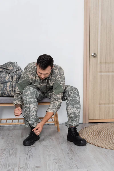 Homem em camuflagem sentado perto da mochila e desatando a bota militar em casa — Fotografia de Stock