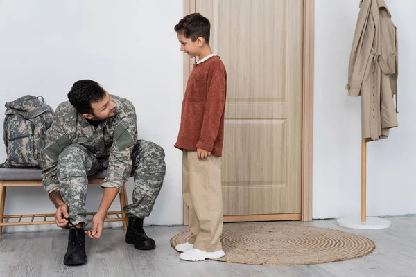 Smiling boy looking at dad in military uniform unlacing boot while sitting near backpack at home — Stock Photo