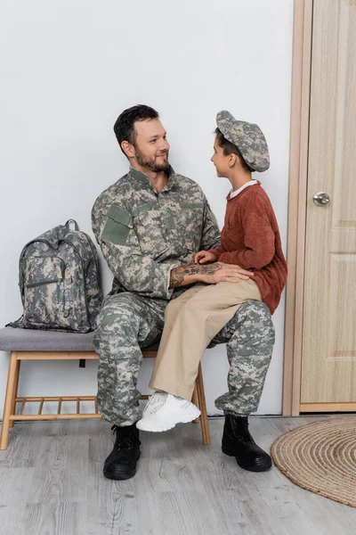 Boy in military cap sitting on laps of dad in camouflage near backpack and entrance door at home — Stock Photo