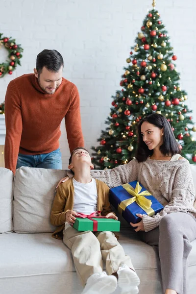 Alegre chico mirando papá mientras sentado en sofá cerca sonriendo mamá y regalos de Navidad - foto de stock