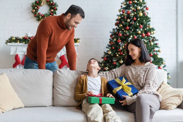Щасливий хлопчик дивиться на тата, сидячи на дивані з різдвяними подарунками і усміхненою мамою — стокове фото