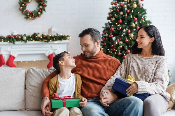 Hombre feliz cogido de la mano con la esposa y mirando al hijo alegre en el sofá cerca de cajas de regalo y el árbol de Navidad borrosa - foto de stock