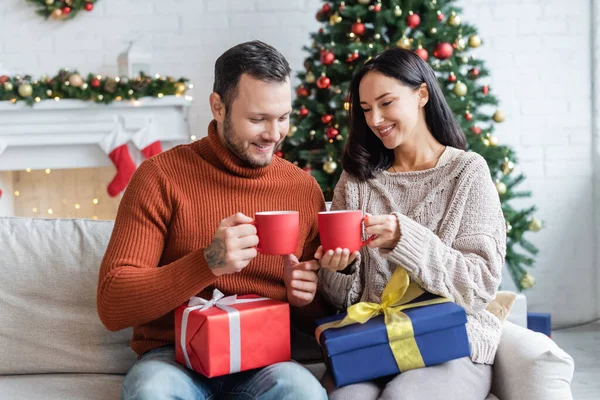 Cheerful couple with gift boxes and cups of warm cocoa sitting near christmas tree on blurred background — Stock Photo
