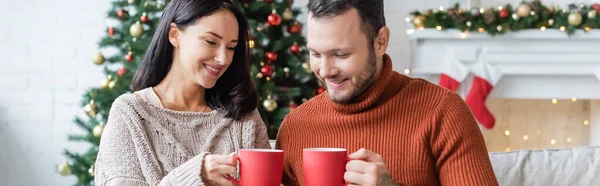 Joyeux couple tenant des tasses de cacao chaud dans le salon avec décor de Noël flou, bannière — Photo de stock