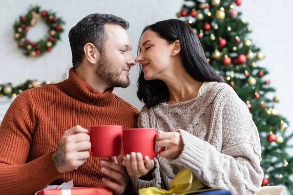 Casal alegre com xícaras de cacau quente sentado cara a cara perto da árvore de natal no fundo borrado — Fotografia de Stock