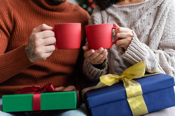 Cropped view of couple holding cups of warm cocoa near christmas presents — Stock Photo