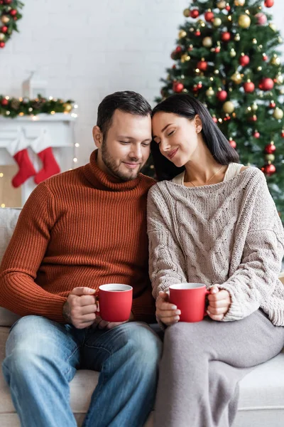 Zufriedenes Paar mit Tassen warmen Kakaos sitzt im Wohnzimmer neben verschwommenem Weihnachtsbaum — Stockfoto