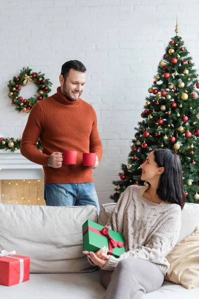 Uomo sorridente in possesso di tazze con cacao caldo vicino moglie felice seduto sul divano vicino regali di Natale — Foto stock