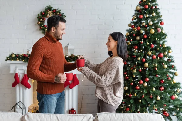 Vista lateral do casal com xícaras de cacau quente sorrindo um para o outro perto da árvore de natal decorada — Fotografia de Stock